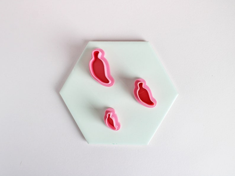 May include: Three pink plastic cookie cutters shaped like chili peppers on a light blue hexagonal surface.