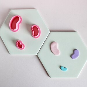 May include: Three pink jelly bean shaped cookie cutters and three jelly beans in pink, purple, and blue on a light green background.