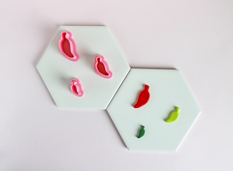 May include: Five different cookie cutters in the shape of chili peppers. The cutters are on a white surface. The cutters are red, green, and pink.