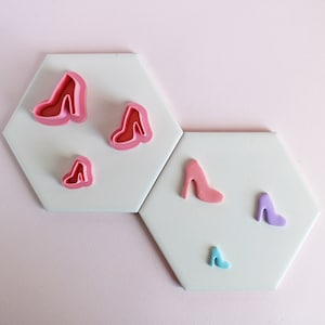 May include: Five different sized cookie cutters shaped like high heels. The cutters are pink and purple and are on a white surface.