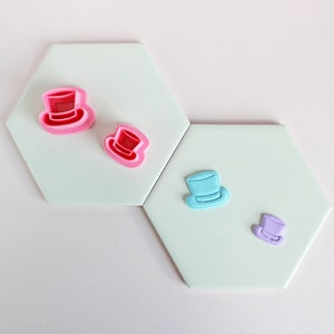 May include: Four different sized cookie cutters in the shape of a top hat. The cutters are pink, light blue, and purple. The cutters are on a light green background.