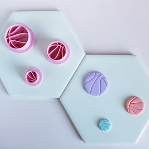 May include: A set of five pink and purple fondant cutters and molds shaped like basketballs. The cutters are different sizes and are arranged on a white surface.