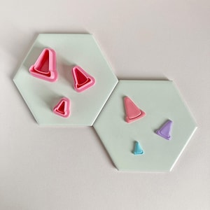 May include: Several cone-shaped cookie cutters in pink and red are displayed on a white hexagonal tile. Another tile features a pink, purple, and blue cutter. These cutters are likely for baking or crafting, with metric measurements.