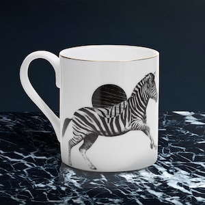 May include: White ceramic mug with a black and white zebra illustration. The mug has a gold rim and a curved handle. The zebra is depicted in mid-stride against a black circle. The mug is on a dark marble surface.