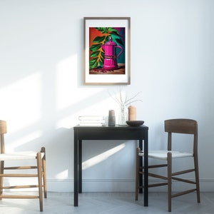 May include: A framed art print featuring a vibrant pink coffee maker with green leaves and coffee beans. The artwork has a colorful background of red, orange, and teal. The print is displayed on a white wall above a black table.