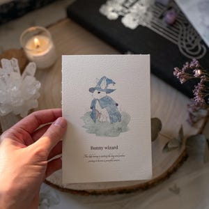 Peut inclure: Un lapin blanc portant un chapeau et une robe de sorcier bleus. La carte indique "Bunny wizard" et "The little bunny is starting the long and powerful journey to become a powerful sorceress."