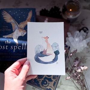 Peut inclure: Une peinture à l'aquarelle d'un renard blanc assis dans un décor de forêt. Le renard regarde un papillon qui vole au-dessus de lui. La peinture est sur une carte blanche avec une bordure bleue.