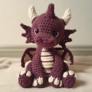 Puede incluir: Un dragón de ganchillo hecho a mano en tonos morados y crema. El dragón tiene ojos grandes y negros, cuernos y alas de color crema. El vientre del dragón presenta rayas crema y moradas. El dragón está sentado sobre una superficie suave de color neutro.