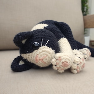 May include: A crocheted stuffed cat toy featuring black and cream yarn. The cat has a black body, cream-coloured paws with pink stitched toe pads, a cream muzzle with pink stitched nose and mouth, and large black plastic safety eyes.