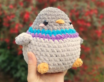 Puede incluir: Un juguete de paloma de ganchillo, redondo y afelpado, hecho de hilo gris suave. Presenta una banda vibrante de hilo azul brillante y morado alrededor del cuello, un pequeño pico amarillo, dos diminutas patas amarillas y ojos de plástico negro.