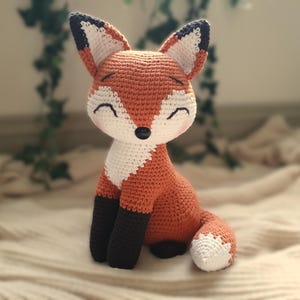 May include: A crocheted fox plush toy with orange and white fur, black eyes, and a black nose. The fox is sitting on a white blanket.