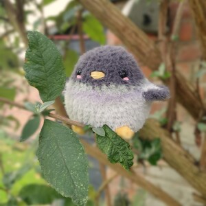May include: A soft, round bird toy in shades of grey, white, and lavender, sitting on a leafy branch. The bird has a yellow beak, pink cheeks, and small wings. The background is blurred, showing tree branches and leaves.