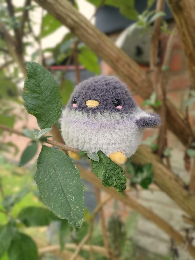 May include: A soft, round bird toy in shades of grey, white, and lavender, sitting on a leafy branch. The bird has a yellow beak, pink cheeks, and small wings. The background is blurred, showing tree branches and leaves.