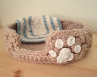 May include: A brown crocheted pet bed with a white paw print design. The bed has a removable blue and white striped liner.