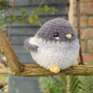 May include: A handmade plush bird toy with a round body, perched on a branch. The bird has gray, white, and lavender fur. It has small black eyes, pink cheeks, and yellow feet. The toy is designed to look like a bird.