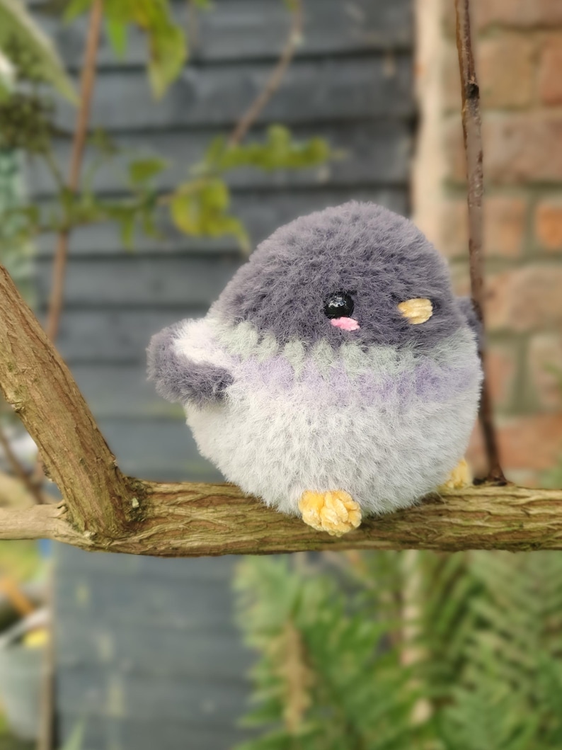 May include: A handmade plush bird toy with a round body, perched on a branch. The bird has gray, white, and lavender fur. It has small black eyes, pink cheeks, and yellow feet. The toy is designed to look like a bird.