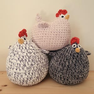 May include: Three crocheted plush chickens in pink, grey, and white. The chickens have red combs and black eyes.