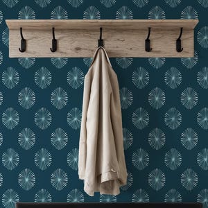 Op de afbeelding: Een marineblauwe muur met een herhalend patroon van witte en lichtblauwe radiale ontwerpen. Een houten plank met zwarte haken is aan de muur bevestigd, met een beige jas die aan een haak hangt. Een zwarte bank staat eronder.