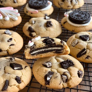 May include: A close-up of a variety of cookies, some with chocolate chips and some with Oreo cookies. The cookies are on a black wire rack.