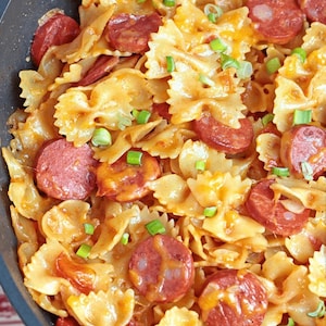 Könnte beinhalten: Eine Nahaufnahme einer Bratpfanne gefüllt mit Schleifenpasta, geschnittener Wurst und geschmolzenem Käse. Die Pasta ist mit gehackten Frühlingszwiebeln bestreut.