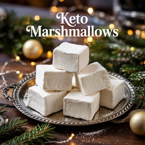 May include: A silver platter with a pile of white keto marshmallows on top. The marshmallows are arranged in a pyramid shape. The platter is surrounded by a garland of lights and a green pine branch.