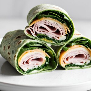 May include: Three green spinach wraps filled with ham and cheese on a white plate.