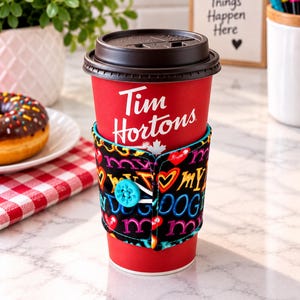 Peut inclure: Un gobelet à café jetable rouge Tim Hortons avec un couvercle en plastique noir, entouré d'une pochette en tissu noir ornée d'un motif coloré de cœurs et du texte "my dog". La pochette est fixée par un bouton bleu vif et une boucle élastique.