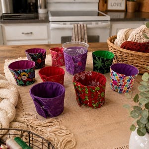 Puede incluir: Una colección de fundas de tela para tazas en varios colores y diseños, incluyendo diseños florales, de corazón y de hojas de cannabis. Las fundas se exhiben sobre una mesa de madera, mostrando sus diversos estilos y texturas. El interior de las fundas es de color sólido.