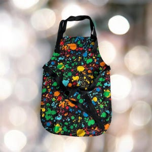 May include: Black apron with a colourful paint splatter pattern. The apron has adjustable straps and a neck strap.