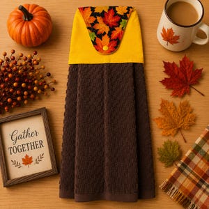 May include: A brown waffle-weave hanging kitchen towel featuring a yellow top panel with a black fabric accent printed with colourful autumn maple leaves and a single yellow snap button closure, centred vertically on a light wood-toned surface.