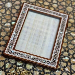Peut inclure: Un cadre photo en bois avec une bordure décorative en nacre et un fond brun clair. Le cadre est posé sur une surface à motif géométrique.