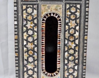 May include: A wooden tissue box cover with intricate inlaid mother-of-pearl designs in black and white. The box has a rectangular shape with a rectangular opening in the front.