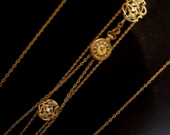 May include: Gold-toned necklace with multiple strands and decorative elements. The necklace features three circular pendants with intricate designs and a small, round watch-like charm. The background is a solid black, highlighting the jewelry's details.