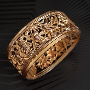 Puede incluir: Un brazalete dorado y ornamentado. El brazalete presenta intrincados diseños calados florales y foliares. El metal tiene un acabado pulido. El diseño es detallado y elegante. El brazalete parece ser de un metal precioso.