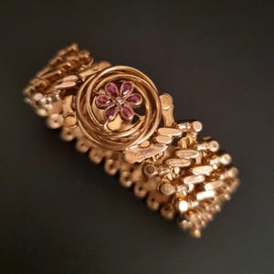 Könnte beinhalten: Ein goldfarbenes Armband mit floralem Mittelstück. Das Armband hat eine Reihe von strukturierten, rechteckigen Gliedern. Das Mittelstück hat ein Spiraldesign mit einer kleinen, roten Blume im Kern. Das Armband ist vor einem dunklen Hintergrund platziert.