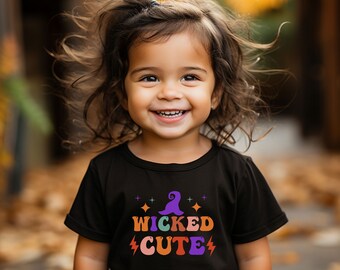 Könnte beinhalten: Ein kleines Kind trägt ein schwarzes T-Shirt mit einem bunten Aufdruck, auf dem "Wicked Cute" und ein Hexenhut zu sehen sind. Das Kind lächelt und hat braunes Haar.