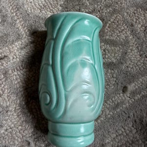 May include: A light mint green ceramic vase featuring a glossy finish and a raised, swirling leaf-like pattern that wraps around the body. The vase has a wide, open top and a distinct, rounded base section with horizontal ridges.