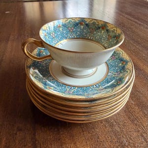 May include: A stack of six matching teacups and saucers. The teacups and saucers are white with a gold rim and a blue and gold floral design. The teacup has a gold handle.