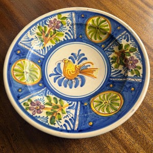 May include: A hand-painted ceramic plate with a white rim and a blue border. The plate features a central design of a yellow bird and blue foliage, surrounded by floral and fruit motifs. The plate measures approximately 10 inches in diameter.