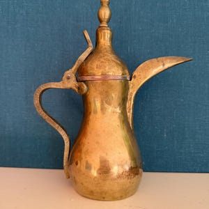 May include: A vintage brass coffee pot with a long spout and a curved handle. The pot has a bulbous body and a decorative finial on the lid. The pot is set against a blue background.