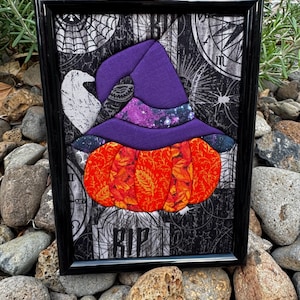 Könnte beinhalten: Ein gerahmtes Stoffkunstwerk mit einem leuchtend orangefarbenen Kürbis, der einen violetten Hexenhut trägt. Der Hintergrund besteht aus schwarz-weißem Stoff mit Spinnennetz- und Uhrenmustern, und die Buchstaben RIP sind unten zu sehen.