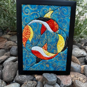 Pode incluir: Uma peça de arte em tecido emoldurada apresentando uma composição circular de dois peixes koi estilizados em laranja, amarelo e branco, sobre um fundo azul vibrante com padrões de redemoinhos dourados e motivos de folhas, em uma moldura preta.