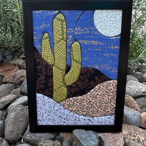 May include: A framed fabric art piece featuring a desert landscape with a green cactus, a blue sky with a white moon, and layered hills in brown and tan floral patterns. The composition uses various textured textiles to create a layered scene.