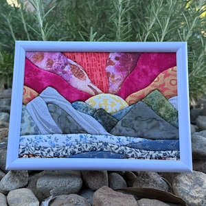 May include: A framed fabric art piece featuring a landscape scene with layered textile mountains in shades of gray and green, a yellow patterned sun, and a vibrant pink and red sky. The foreground consists of blue floral patterned fabric strips.