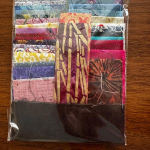 May include: A clear plastic bag containing a collection of assorted fabric squares and strips in various colors and patterns, including floral, geometric, and solid designs, stacked and arranged in a rectangular bundle for sewing or quilting projects.