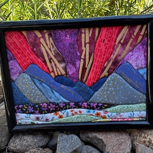 May include: A rectangular fabric art piece in a black frame featuring a landscape scene. The composition includes layered blue mountain shapes, a vibrant purple and red sky with gold accents, and a foreground of floral patterned fabric strips.
