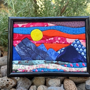 May include: A framed fabric collage art piece featuring a landscape with dark blue mountains, a bright yellow sun, and layered horizontal bands of colorful patterned textiles in shades of red, orange, blue, and floral prints within a black frame.