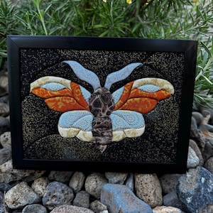 Pode incluir: Uma peça de arte têxtil emoldurada apresentando uma mariposa com asas feitas de tecido estampado em laranja, azul claro e creme. A mariposa tem um corpo texturizado castanho escuro e antenas azul claro sobre um fundo preto com salpicos dourados.