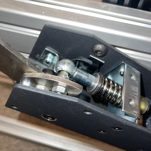 Op de afbeelding: Close-up van een zwart metalen voetpedaal met een zilveren veer en een zilveren kogelgewricht. Het pedaal is bevestigd aan een zilveren metalen frame.