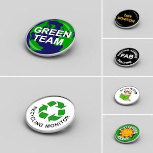 May include: Assortment of colourful round badges with various messages. The badges feature a green recycling symbol, a green earth, a smiling sun, a bookworm, and a black badge with the words "Tidy Monitor".